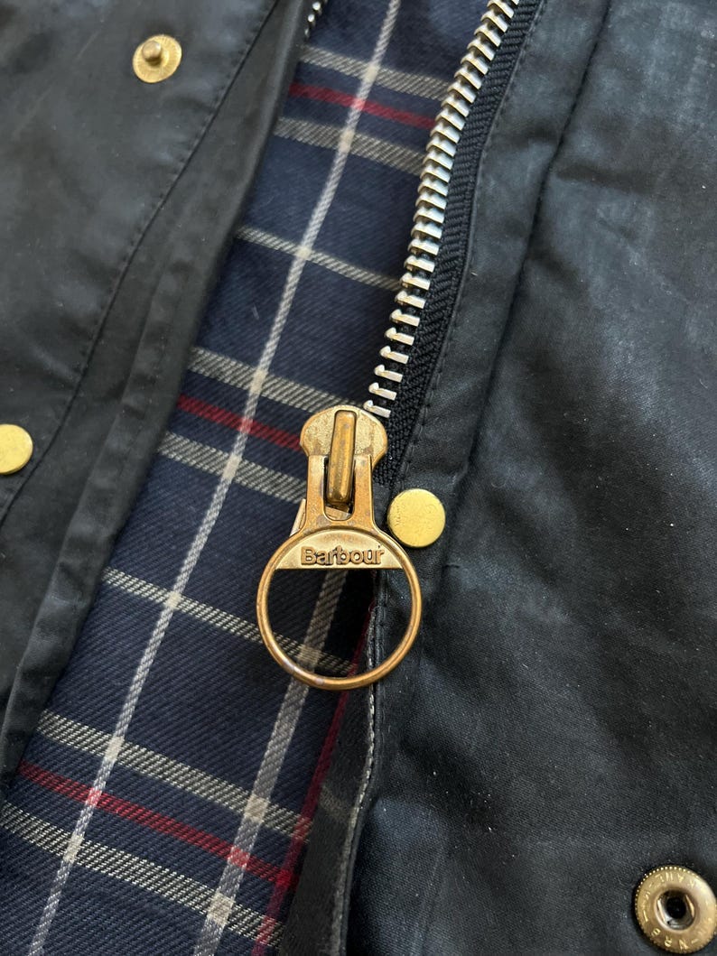 Peut inclure: Gros plan sur une fermeture &eacute;clair de veste Barbour noire avec un tirette en laiton sur laquelle est inscrit "Barbour". La fermeture &eacute;clair est partiellement ouverte, r&eacute;v&eacute;lant une doublure en tissu &agrave; carreaux bleu et blanc.