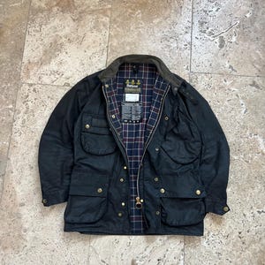 Peut inclure: Une veste Barbour bleu fonc&eacute; avec une doublure &agrave; carreaux et un patch logo Barbour. La veste a plusieurs poches et une fermeture &eacute;clair.