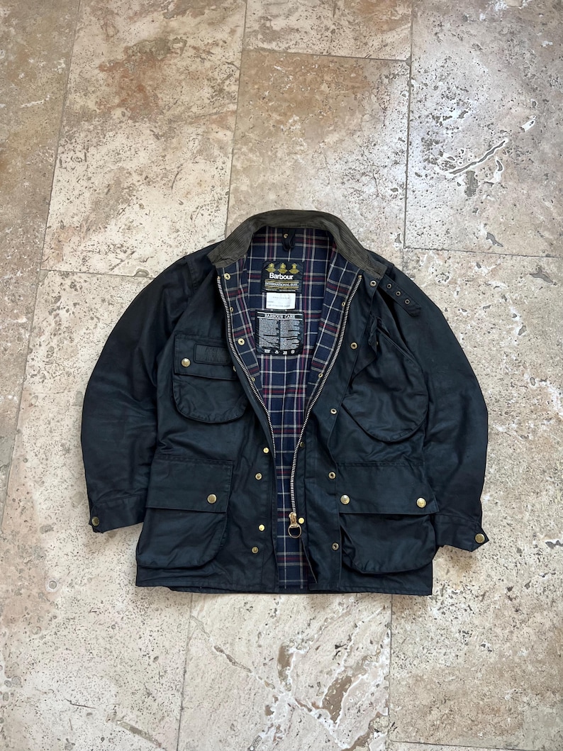 Peut inclure: Une veste Barbour bleu fonc&eacute; avec une doublure &agrave; carreaux et un patch logo Barbour. La veste a plusieurs poches et une fermeture &eacute;clair.