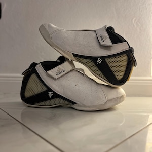 May include: White and black Adidas basketball shoes with a black strap and the Adidas logo on the side. The shoes have a white mesh panel and a black and white logo on the back.