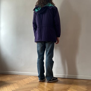 Pu&ograve; includere: Una giacca con cappuccio viola con fodera turchese. La giacca ha una vita con coulisse e viene indossata sopra un paio di jeans blu.