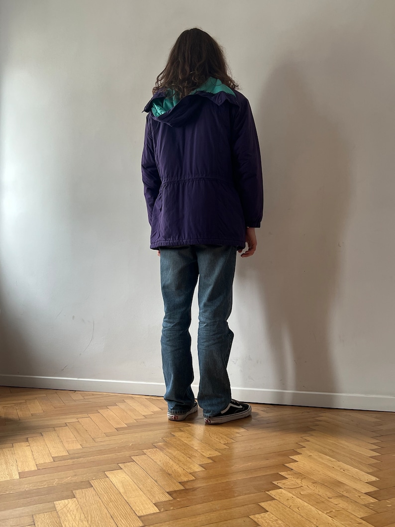 Pu&ograve; includere: Una giacca con cappuccio viola con fodera turchese. La giacca ha una vita con coulisse e viene indossata sopra un paio di jeans blu.
