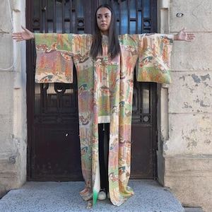 Peut inclure: Un kimono long et orné de motifs floraux dans les tons de vert, rose et jaune. Le vêtement est porté ouvert, révélant un pantalon noir et des chaussures blanches. L'arrière-plan comprend une porte marron foncé et un mur en pierre vieilli.
