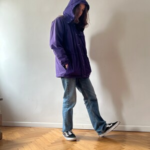 Pu&ograve; includere: Una giacca viola con cappuccio, chiusura a zip e diverse tasche. La giacca &egrave; indossata sopra un paio di jeans blu e scarpe da ginnastica nere.