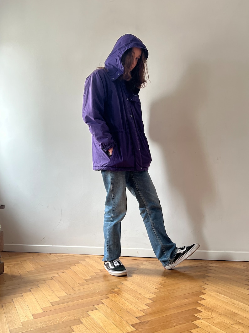 Pu&ograve; includere: Una giacca viola con cappuccio, chiusura a zip e diverse tasche. La giacca &egrave; indossata sopra un paio di jeans blu e scarpe da ginnastica nere.