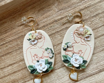 May include: A pair of cream-colored earrings with gold-colored hoops and posts. Each earring features a floral design with a heart-shaped charm. The earrings have a line drawing of a mother and child.
