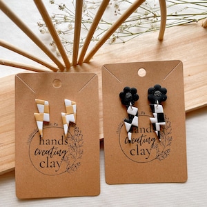 May include: Two pairs of handmade polymer clay earrings. The first pair features black and white checkered lightning bolt shapes. The second pair features black and white checkered lightning bolt shapes with a black flower charm on each earring. Both pairs of earrings are displayed on brown card stock with the text "handmade" and "hands creating clay" on each card.