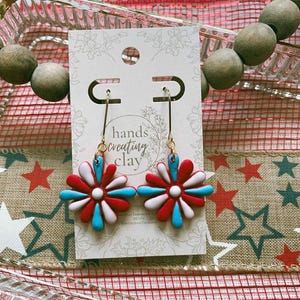 May include: A pair of handmade flower earrings with red, white, and blue petals. The earrings are on a white card with the text "hands creating clay". The earrings hang from gold-colored hooks.