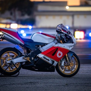 Pode incluir: Uma motocicleta branca, vermelha e preta com rodas douradas. A motocicleta tem a palavra "Bimota" escrita no lado.