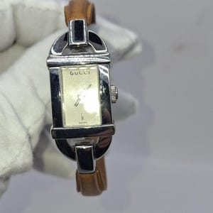 May include: A rectangular Gucci watch with a silver-tone case and a light brown leather strap. The watch face is cream-colored with silver hands and the Gucci logo. The watch is held against a white background.