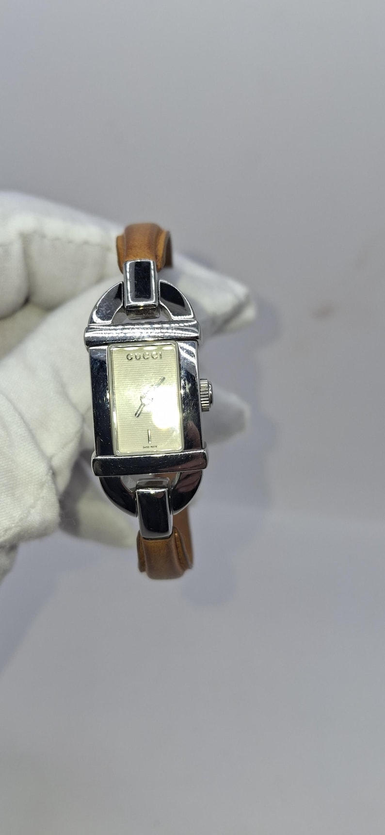 May include: A rectangular Gucci watch with a silver-tone case and a light brown leather strap. The watch face is cream-colored with silver hands and the Gucci logo. The watch is held against a white background.