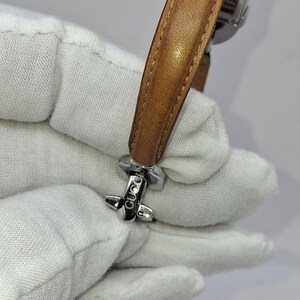 May include: A silver-toned wristwatch with a light brown leather band. The watch has a rectangular face and a buckle clasp. The band is approximately 1.5 cm wide. The watch is held by a gloved hand.