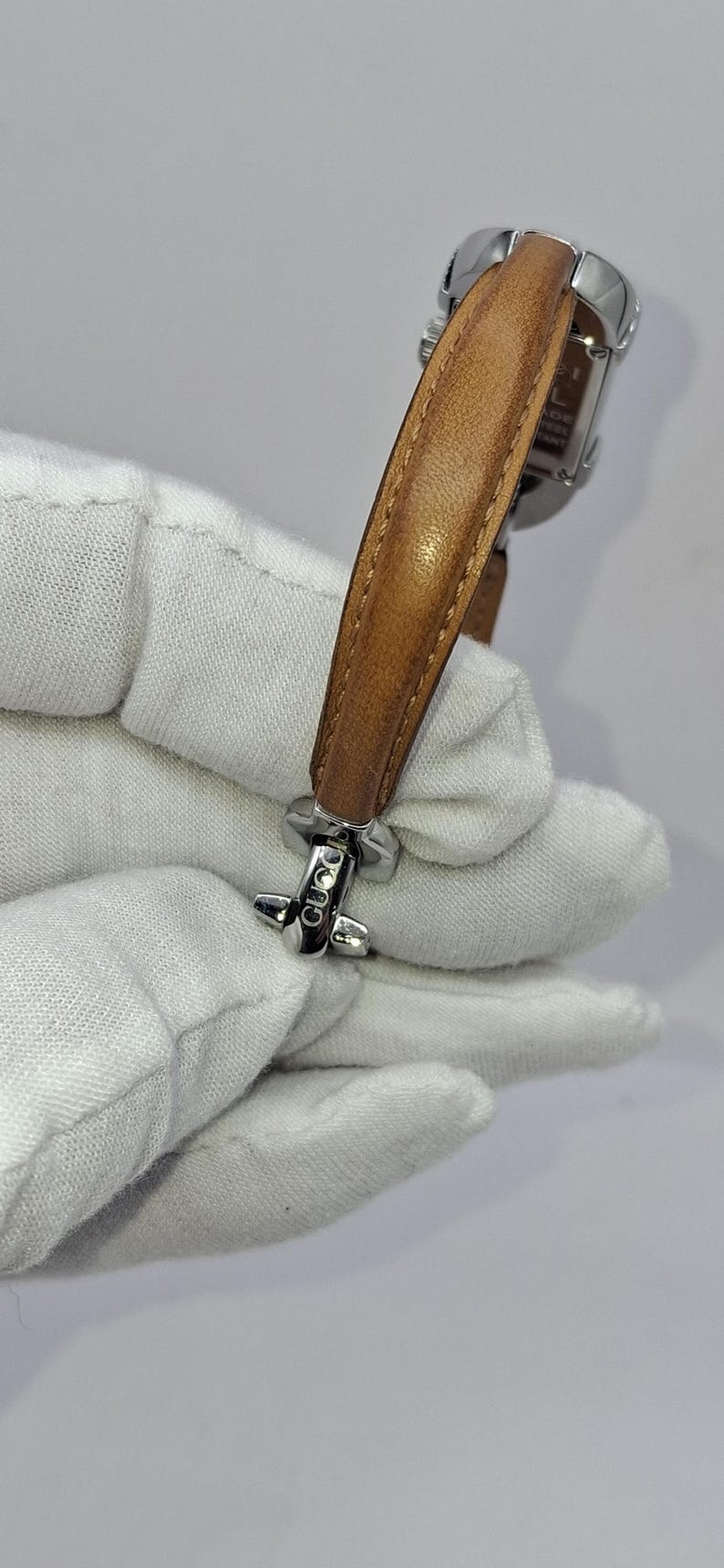 May include: A silver-toned wristwatch with a light brown leather band. The watch has a rectangular face and a buckle clasp. The band is approximately 1.5 cm wide. The watch is held by a gloved hand.