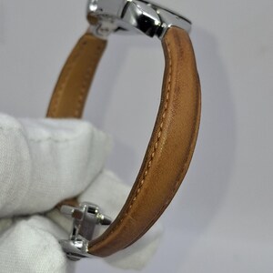 May include: A wristwatch with a light brown leather band and a silver-colored metal clasp. The watch band is curved, and the stitching is visible along the edges. The watch face is not visible.