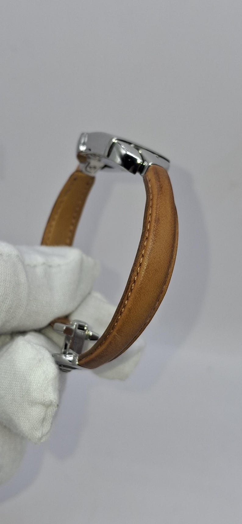 May include: A wristwatch with a light brown leather band and a silver-colored metal clasp. The watch band is curved, and the stitching is visible along the edges. The watch face is not visible.