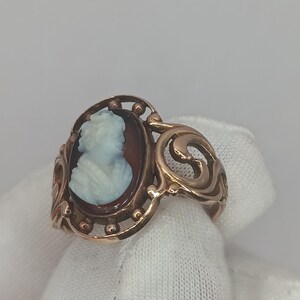 May include: A gold-toned ring featuring a cameo portrait of a woman in profile. The cameo is set against a dark background within an oval frame. The ring has an ornate, swirling design on the sides.