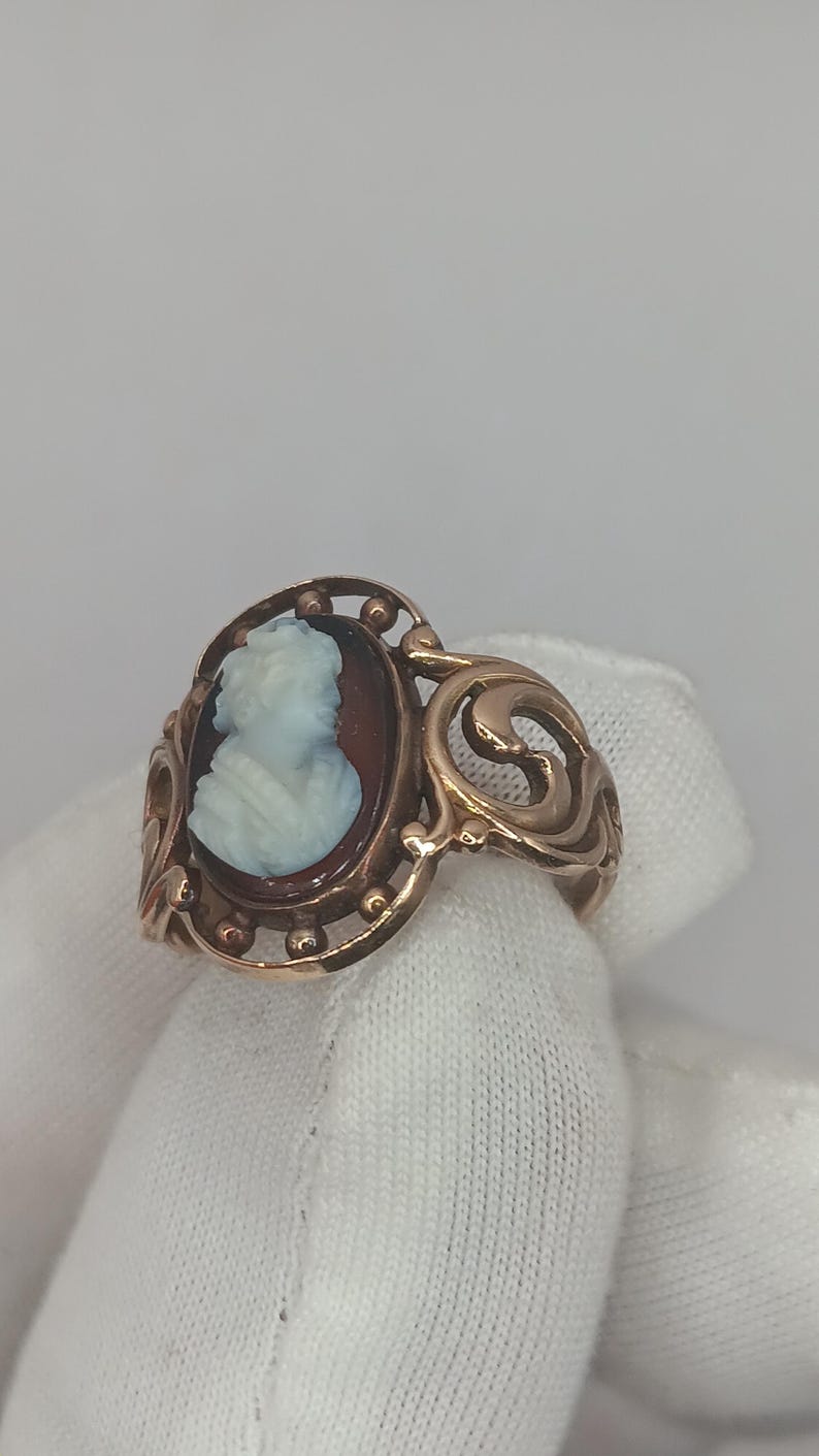 May include: A gold-toned ring featuring a cameo portrait of a woman in profile. The cameo is set against a dark background within an oval frame. The ring has an ornate, swirling design on the sides.