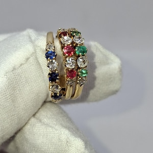 May include: Three gold rings with colorful gemstones. One ring features blue and white stones, another has red, white, and green stones, and the third ring is a simple gold band. The rings are held against a white background.