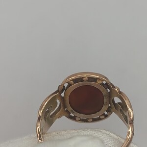 May include: A gold-toned ring featuring an oval, reddish-brown stone. The ring has an ornate, openwork design around the stone and on the shoulders. The ring is set against a white background.
