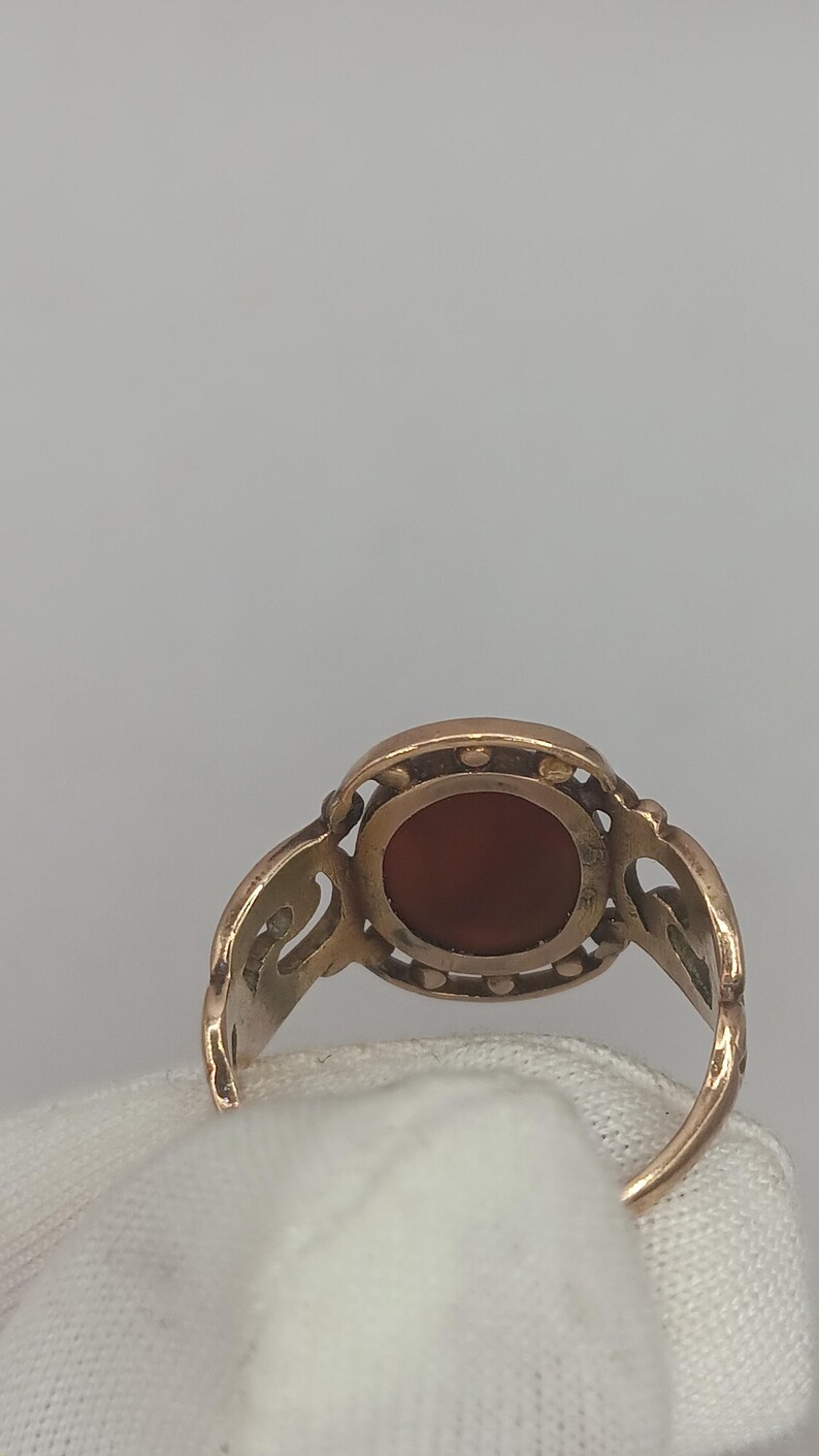 May include: A gold-toned ring featuring an oval, reddish-brown stone. The ring has an ornate, openwork design around the stone and on the shoulders. The ring is set against a white background.