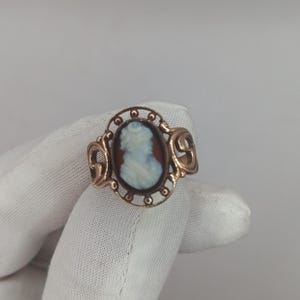 May include: A gold-toned ring featuring a cameo with a profile of a woman in white against a brown background. The ring has an oval setting with decorative filigree and scrollwork on the sides. The ring is held by a gloved hand.
