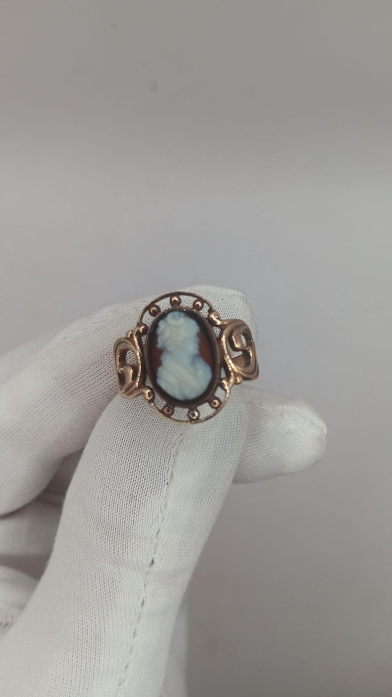 May include: A gold-toned ring featuring a cameo with a profile of a woman in white against a brown background. The ring has an oval setting with decorative filigree and scrollwork on the sides. The ring is held by a gloved hand.