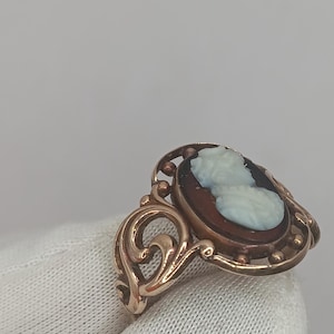 May include: A vintage cameo ring featuring a profile of a woman in white against a dark brown background. The ring has a gold-colored filigree setting with an oval shape and decorative swirls.