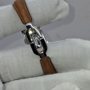 May include: A silver-toned watch with a brown leather strap. The watch has a rectangular face and a sleek, modern design. The strap is made of genuine leather.