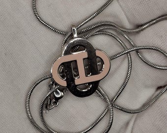 May include: Silver-toned necklace with a pendant featuring a double-circle design with a pink center. The chain is a thin, snake-style design. The pendant has a shiny, reflective surface.