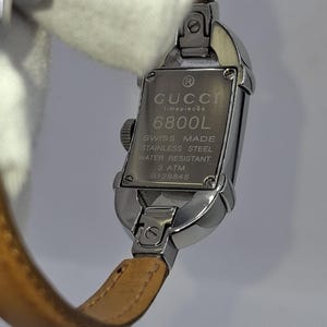 May include: A silver-toned Gucci timepiece with a rectangular watch face and a brown leather strap. The watch face displays the Gucci logo, model number 6800L, and other details. The watch is water-resistant.