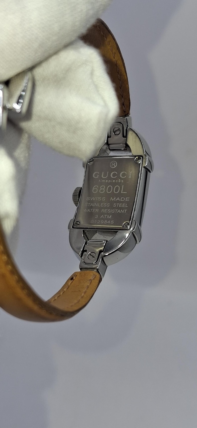 May include: A silver-toned Gucci timepiece with a rectangular watch face and a brown leather strap. The watch face displays the Gucci logo, model number 6800L, and other details. The watch is water-resistant.