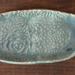 May include: A handcrafted, oval-shaped ceramic serving dish. The light blue dish features embossed floral and leaf patterns. The rim is slightly raised, and the dish has a smooth, glossy finish. The dimensions are approximately 12 inches long and 6 inches wide.