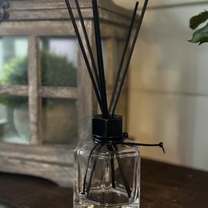 May include: A clear glass diffuser bottle with a black square top and black reeds. The bottle is tied with a black cord and a decorative silver bead. The diffuser sits on a dark wooden surface.