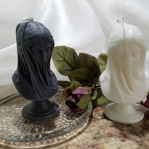 May include: Two bust-shaped candles. One is dark gray, the other is off-white. Both candles feature a draped fabric design, giving them a classical appearance. The candles are on a clear glass plate, with greenery and flowers in the background. These candles are ideal for home decor.