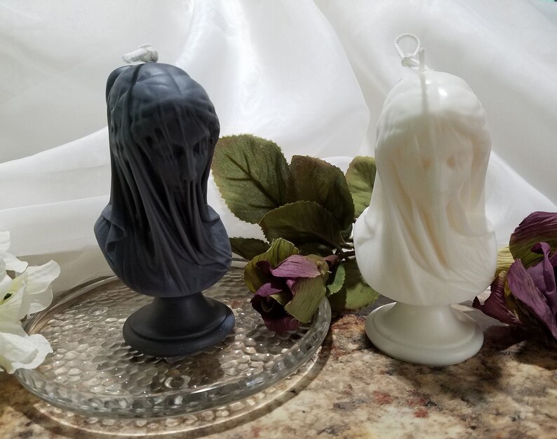 May include: Two bust-shaped candles. One is dark gray, the other is off-white. Both candles feature a draped fabric design, giving them a classical appearance. The candles are on a clear glass plate, with greenery and flowers in the background. These candles are ideal for home decor.