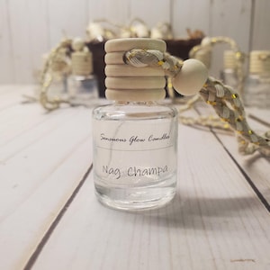 May include: A clear glass bottle with a wooden cap and a braided cord. The bottle contains liquid and has a label that reads "Sensuous Glow Candles" and "Nag Champa". The background includes other bottles and decorative elements.