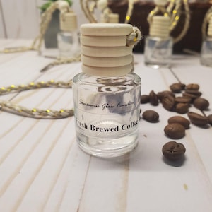 May include: A clear glass bottle with a wooden cap and a string. The bottle has the text "Sensuous Glow Candles Fresh Brewed Coffee". Coffee beans are scattered around the bottle. The bottle is on a light-colored wooden surface.