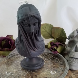 May include: A dark gray candle shaped like a veiled bust, with a wick at the top. A gray skull with a snake wrapped around it is next to the candle. The candle and skull are on a glass plate, with a dark red rose and green leaves in the background.