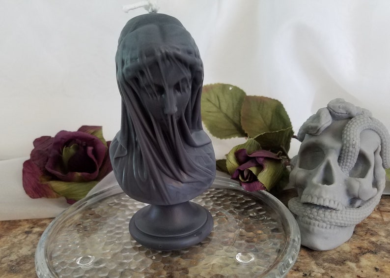 May include: A dark gray candle shaped like a veiled bust, with a wick at the top. A gray skull with a snake wrapped around it is next to the candle. The candle and skull are on a glass plate, with a dark red rose and green leaves in the background.