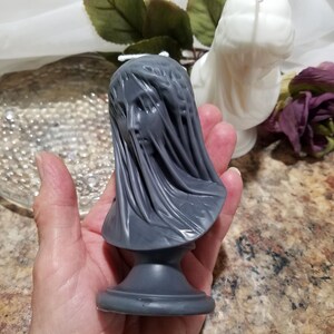 May include: A gray candle shaped like a veiled bust. The candle features a detailed face and draped fabric, mounted on a pedestal base. A white candle and a purple rose are in the background.
