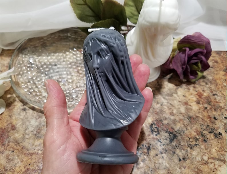 May include: A gray candle shaped like a veiled bust. The candle features a detailed face and draped fabric, mounted on a pedestal base. A white candle and a purple rose are in the background.