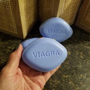 May include: Light blue, oval-shaped soaps with the word "VIAGRA" embossed on each. Several soaps are visible, with one held in a hand. The background includes a dark countertop and woven wooden panels.
