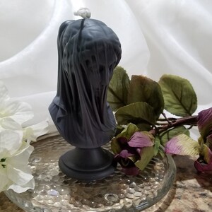 May include: A dark gray candle shaped like a veiled bust, set on a black pedestal. The candle has a wick at the top and is surrounded by white flowers and a purple rose on a glass plate.