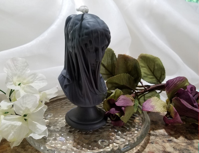 May include: A dark gray candle shaped like a veiled bust, set on a black pedestal. The candle has a wick at the top and is surrounded by white flowers and a purple rose on a glass plate.