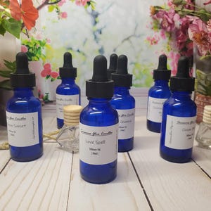 May include: Several cobalt blue glass dropper bottles with black caps and white labels. Each label features the text Sensuous Glow Candles, a scent name such as Love Spell, and the volume 30ml. Small wooden diffuser caps are placed near the bottles.