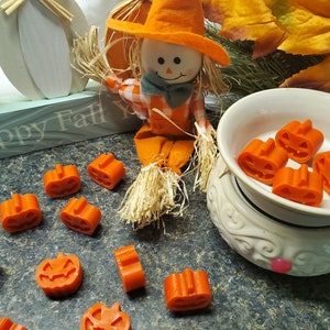 May include: Orange pumpkin-shaped wax melts scattered around a decorative wax warmer. A scarecrow figurine and a wooden sign that says "Happy Fall Y'all" are in the background. The wax melts are approximately 1 inch in size.