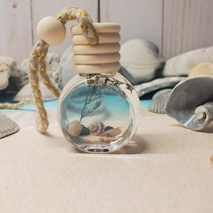 May include: A round, clear glass bottle filled with a light blue liquid, small seashells, and a dried plant sprig. The bottle features a ribbed wooden cap and a thick, beige braided hanging cord with a single wooden bead attached to the top.