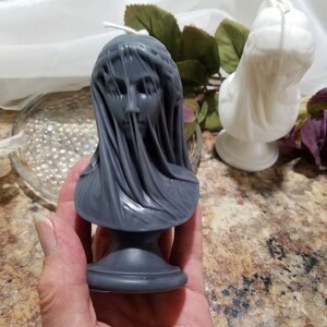 May include: A gray candle shaped like a bust of a veiled woman. The candle has a detailed face and draped fabric. A white candle in a similar style is in the background. The candles are on a brown surface.