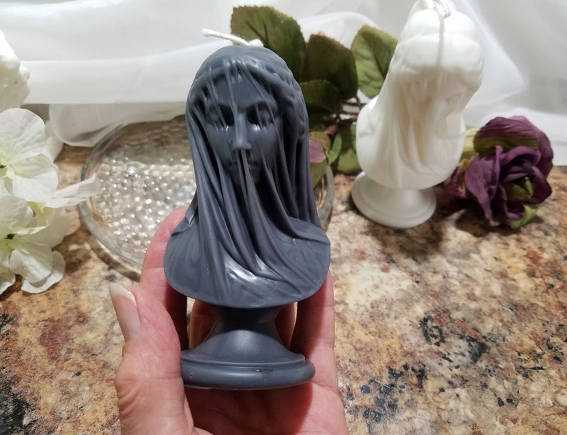 May include: A gray candle shaped like a bust of a veiled woman. The candle has a detailed face and draped fabric. A white candle in a similar style is in the background. The candles are on a brown surface.
