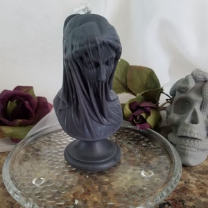May include: A dark gray candle shaped like a veiled bust, set on a glass plate. A gray skull with snakes and a burgundy rose are also visible. The candle bust has a draped veil over the face.